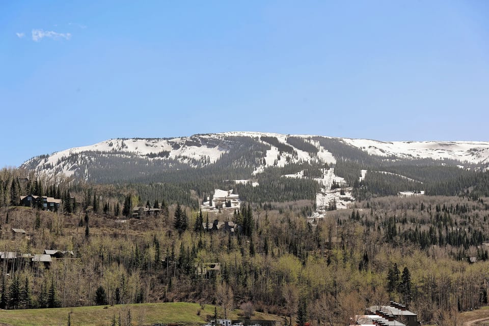 A mountain landscape showing a snow-covered ridge, partially forested slopes, and scattered buildings nestled among trees under a clear blue sky.