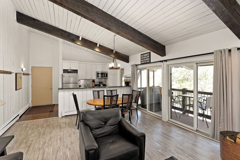 Spacious living area with vaulted ceiling, exposed beams, open kitchen with white cabinets, stainless steel appliances, round dining table with six chairs, leather armchairs, and sliding glass doors to a balcony.