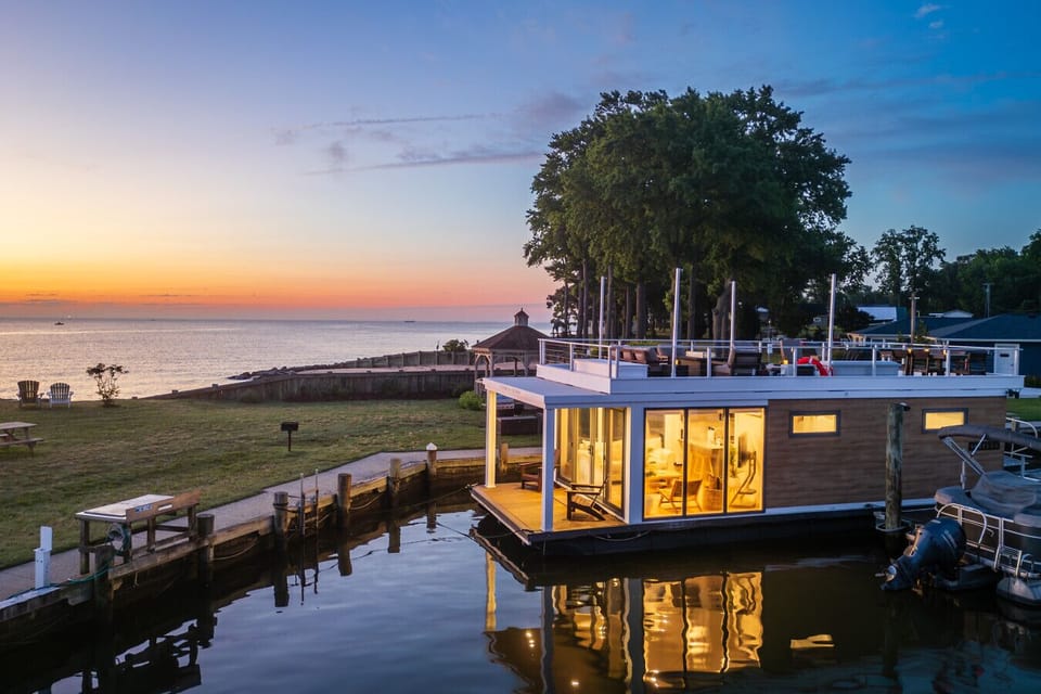 Twilight exterior view of the floating home glowing from within, framed by tall trees and the Chesapeake Bay`s last orange light behind.