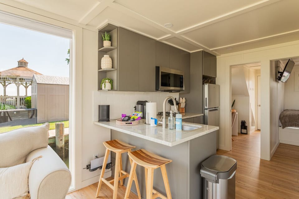 Compact modern kitchen with gray shaker cabinetry, white subway tile, Nespresso coffee maker, two-burner gas cooktop, and a breakfast bar looking out to the marina gazebo.