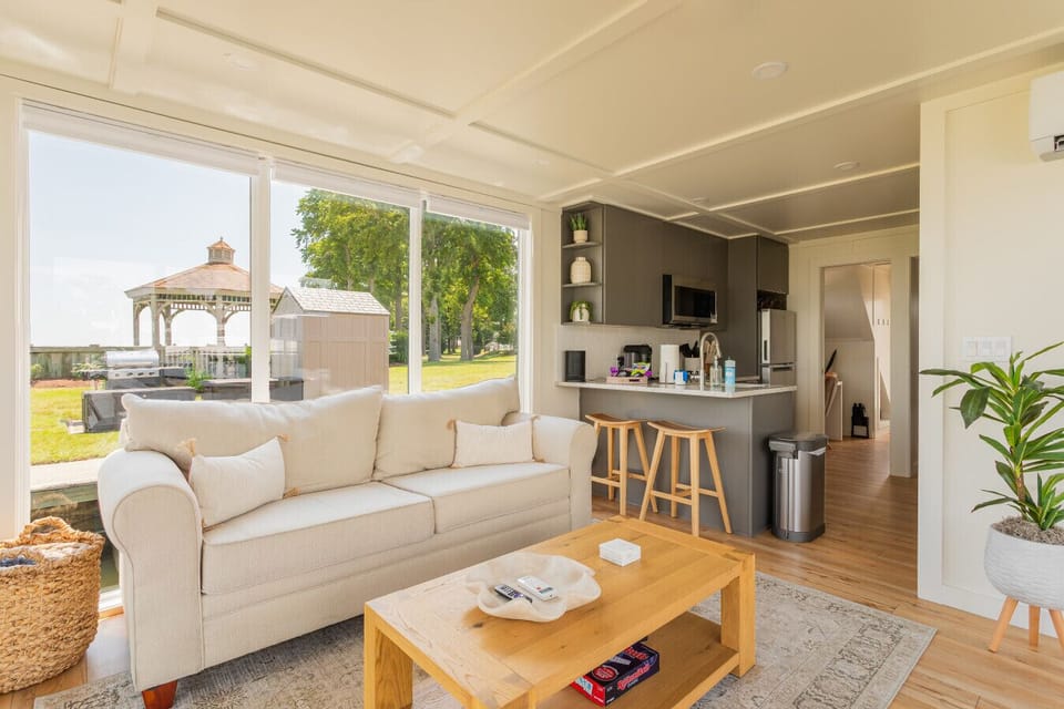 Main salon styled with a linen loveseat, oak coffee table, and picture windows framing the shoreside gazebo and picnic-grill area of Patapsco Marina.