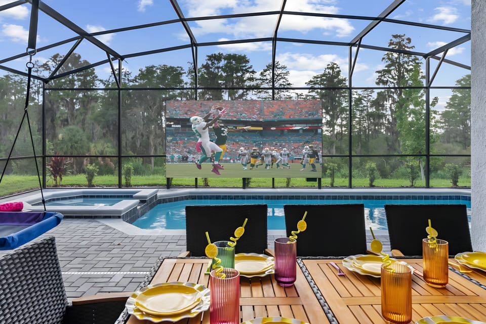 Enjoy our vibrant outdoor dining area with a beautiful wooden table and colorful glassware, perfect for al fresco meals. Relax by the glistening pool under the open sky, complete with a large screen for entertainment.