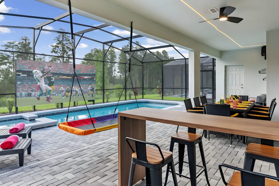 Relax in a vibrant outdoor space with a colorful swing by the shimmering pool. Enjoy modern bar seating and a sleek dining table, all nestled among lush greenery in a comfortable screened enclosure.