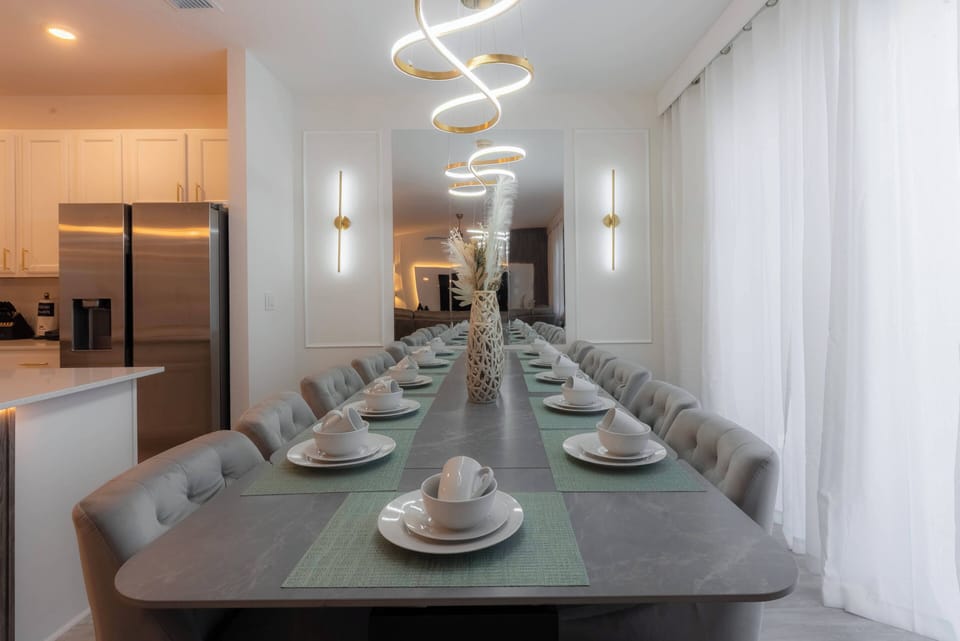 Enjoy communal dining in a beautiful space with a sleek elongated table and tasteful tableware. The soft glow of modern pendant lighting enhances the atmosphere, complemented by large windows dressed in light, flowing curtains.