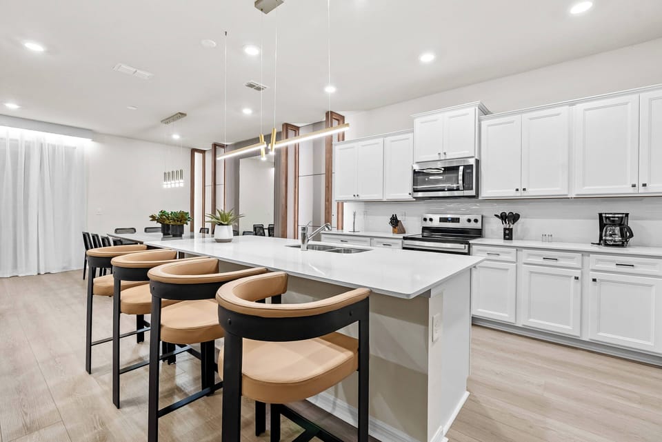 The bright kitchen features sleek white cabinetry and a spacious island with stylish light wood stools, creating a welcoming atmosphere enhanced by warm overhead lighting and large windows that fill the space with natural light.