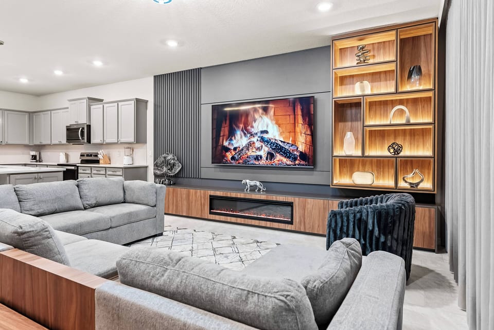 Relax in this welcoming living room featuring a stunning wall-mounted fireplace. Enjoy the stylish gray sofas and charming decor on wooden shelves, all enhanced by the soft, inviting glow of ambient lighting.