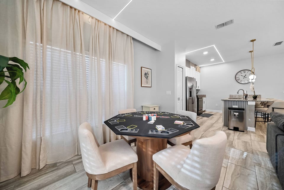 Sunlight floods the stylish dining area, highlighting the unique octagonal table and plush white chairs. Soft beige curtains and light wood flooring add warmth, creating an inviting space for meals and conversations.