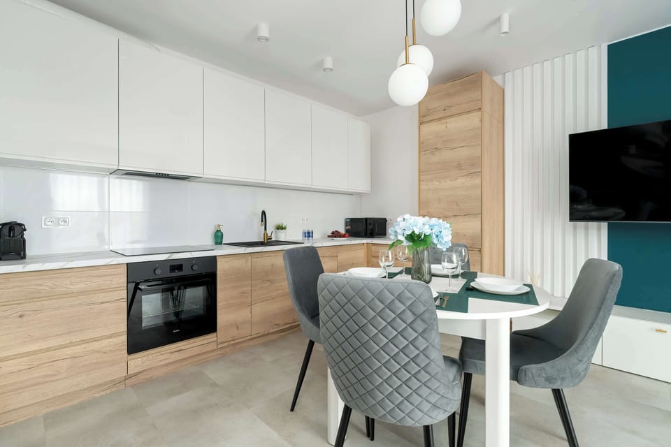 The picture shows a bright kitchen with white cabinets and a wooden worktop, open to a dining area with a round table and grey quilted chairs. 