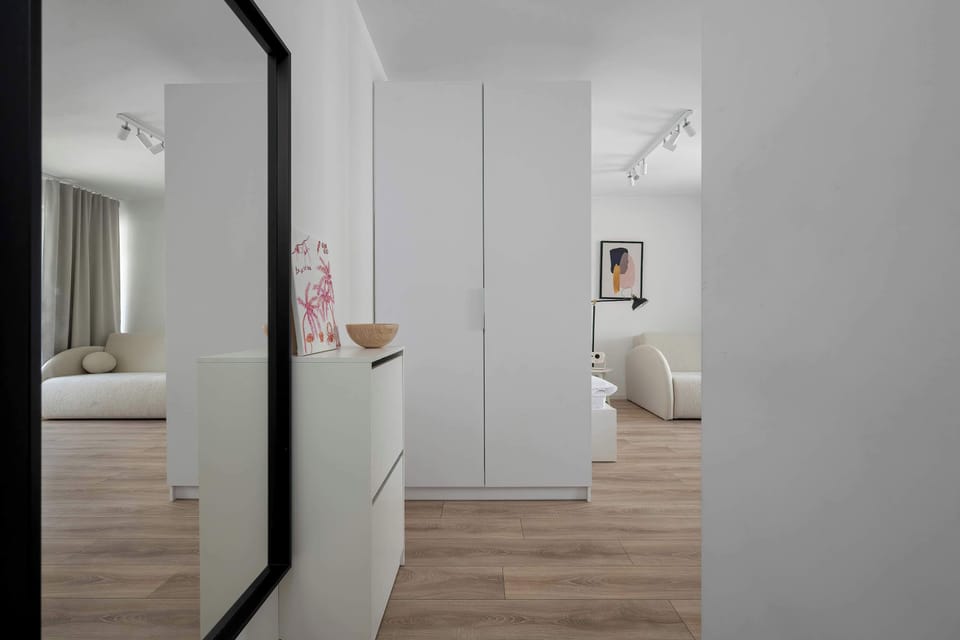 A mirror in the hallway reflects the bright interior of the studio, showing the white wardrobe and a glimpse of the living area. The light wood flooring adds warmth to the clean, white space.