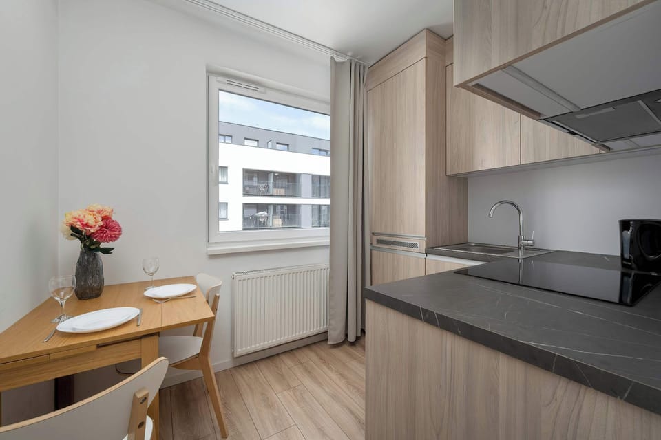 A view of the kitchenette highlights a small wooden dining table positioned near a window. The dark grey countertops and light wood cabinets provide a modern contrast in the bright culinary space.