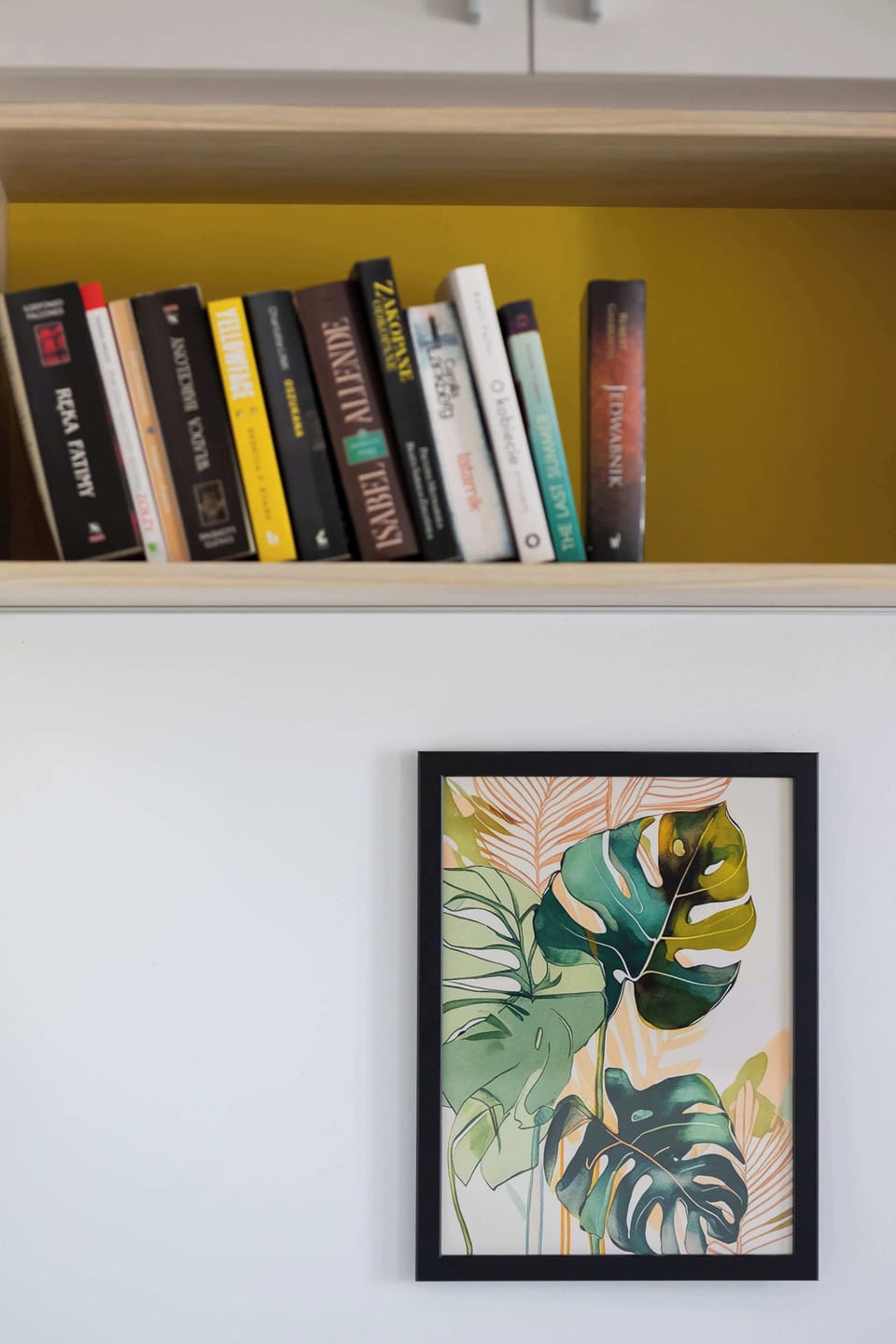 Discover a touch of culture and art, where a shelf lined with diverse books sits above a vibrant tropical leaf print, adding character and charm to the living space.