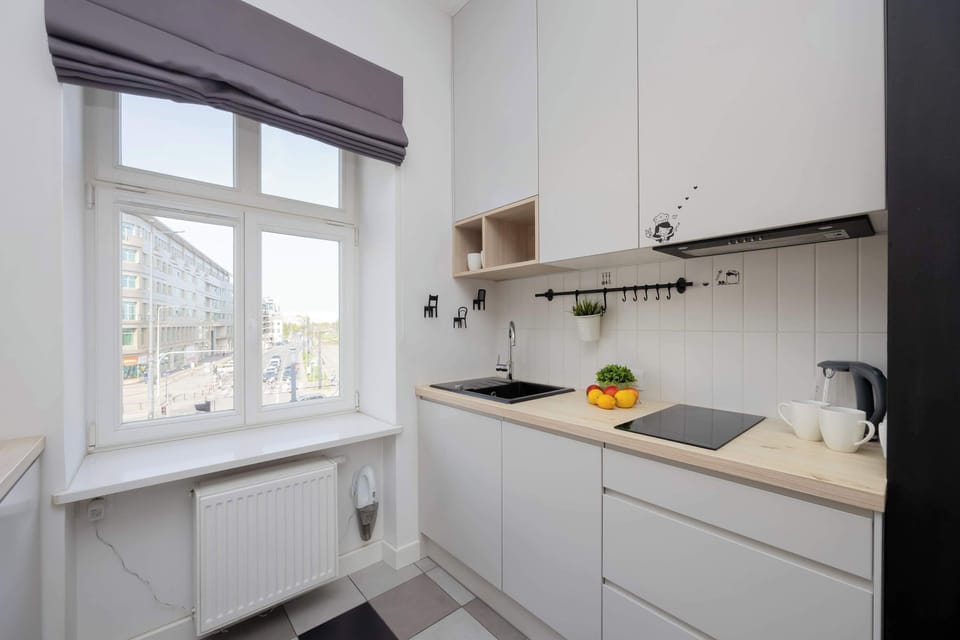 Discover a bright and well-equipped kitchenette in this Gdańsk studio, featuring modern white cabinets, a sleek black sink, and essential appliances, all situated by a window offering a view of the city.