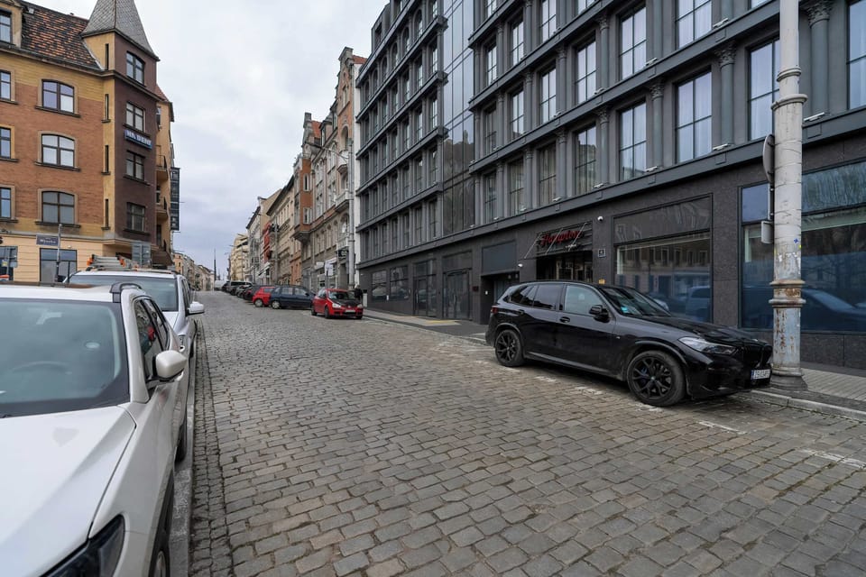 A perspective down the cobblestone street, lined with parked cars and a mix of historic and contemporary architecture.






