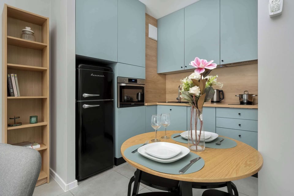 A compact yet functional kitchen area with open shelving, full-size refrigerator, and countertop with dining setup.

