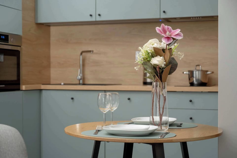 A modern kitchen with sleek light blue cabinetry, wooden accents, and a black built-in oven, complemented by a round dining table set for two with fresh flowers, creating a cozy and stylish dining space.