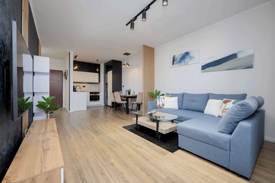 A modern living room with a blue sofa, coffee table, and a large window. The room is decorated with houseplants and minimalistic art, giving it a cozy yet contemporary feel.