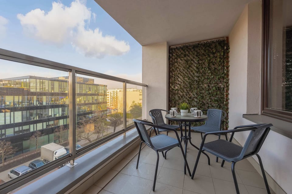 A private balcony featuring a small table and chairs, perfect for relaxing while enjoying an open view of modern architecture and green spaces.