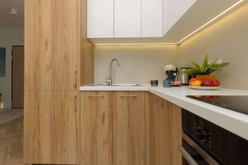 A minimalist kitchen featuring wooden lower cabinets, white upper cabinets, and a black countertop. Small potted plants and kitchen essentials add charm.