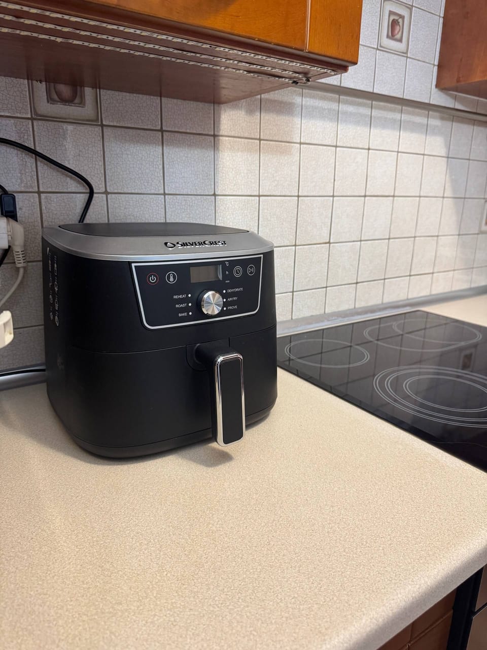 A black air fryer stands on the kitchen counter near the stove. White tiles and wooden cabinets can be seen in the background.