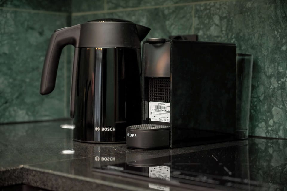 A close-up of a kitchen corner with a black electric kettle and matching appliances. The green backsplash and wood countertop complement the aesthetic.