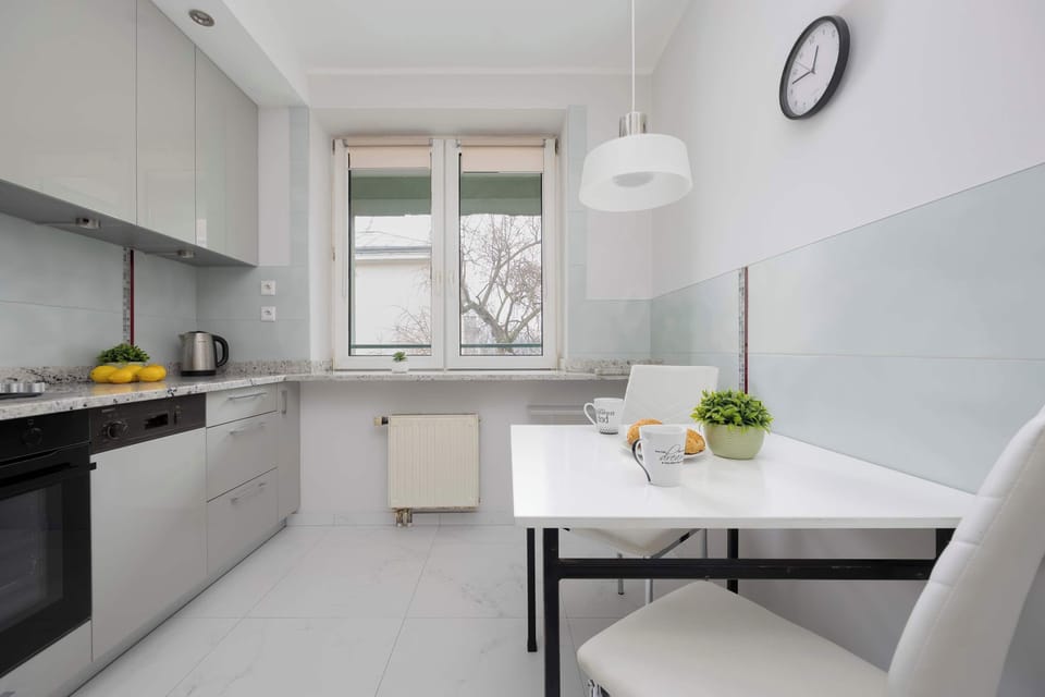 A minimalist kitchen with a white dining table, a black clock on the wall, and a window.