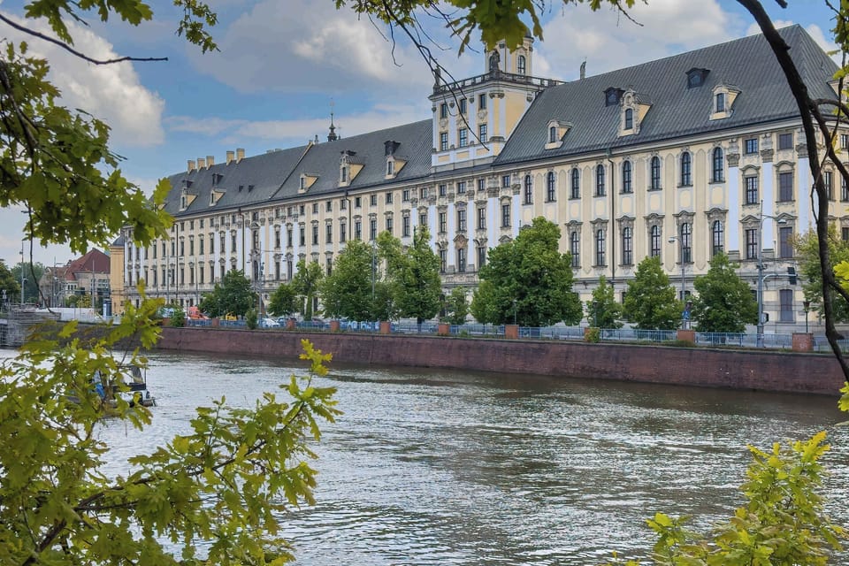 A scenic riverfront view with a historic Baroque-style building on the opposite bank, framed by tree branches in the foreground.