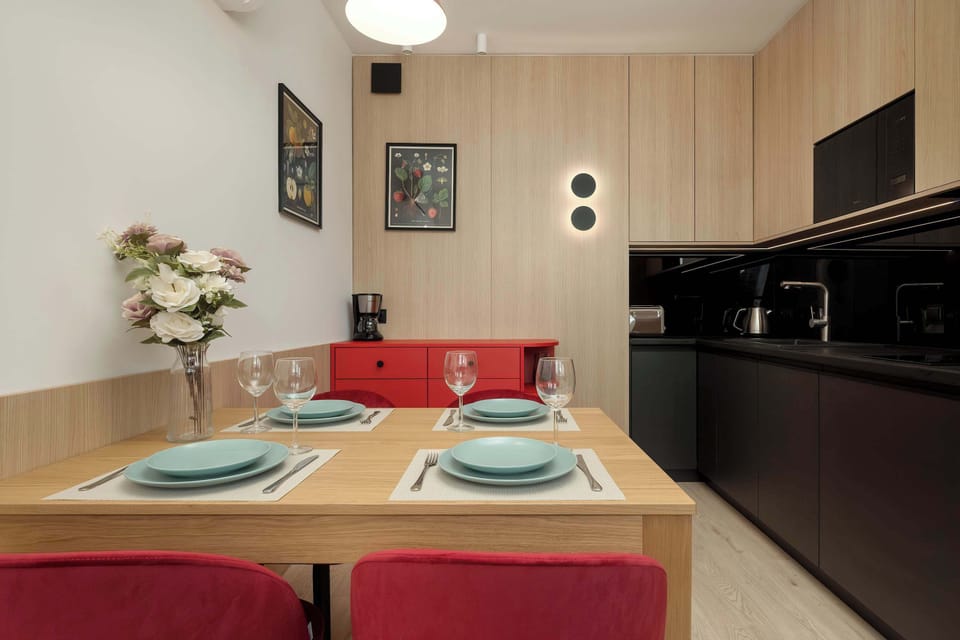 A neatly set dining area for four with matching dishware and deep red chairs, with a view toward the open-plan kitchen.