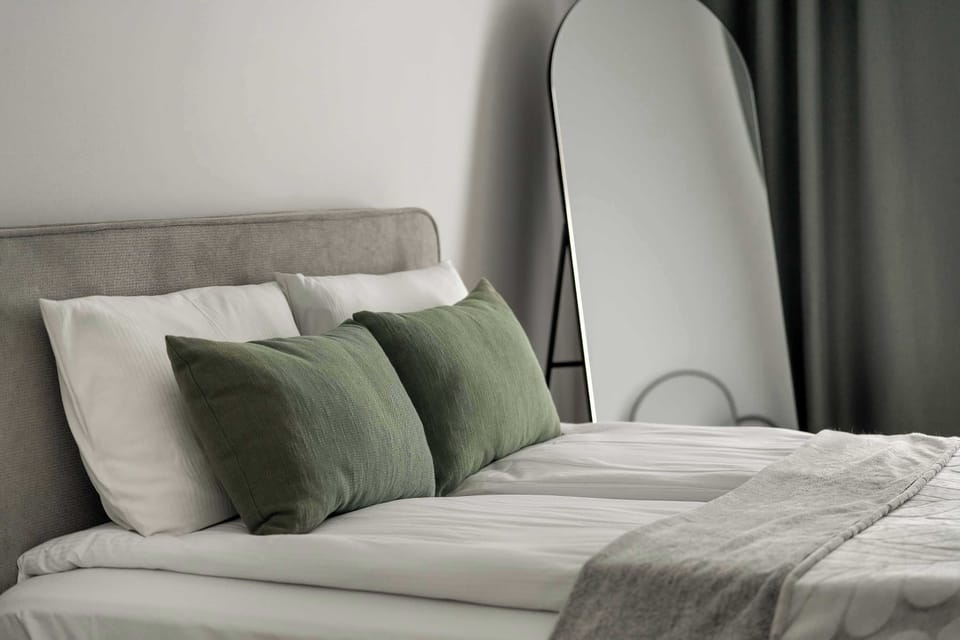 A bright bedroom featuring a bed with soft green cushions and modern bedside lighting. The space is airy, calm, and inviting.
