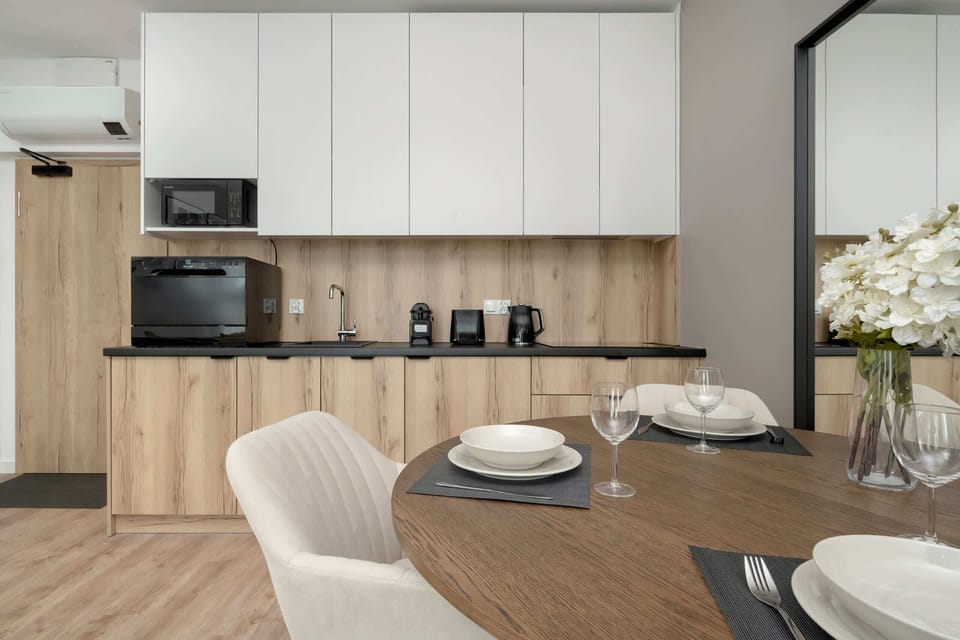 A kitchenette featuring light wood and white cabinetry, black appliances, and a dining table with beige chairs.