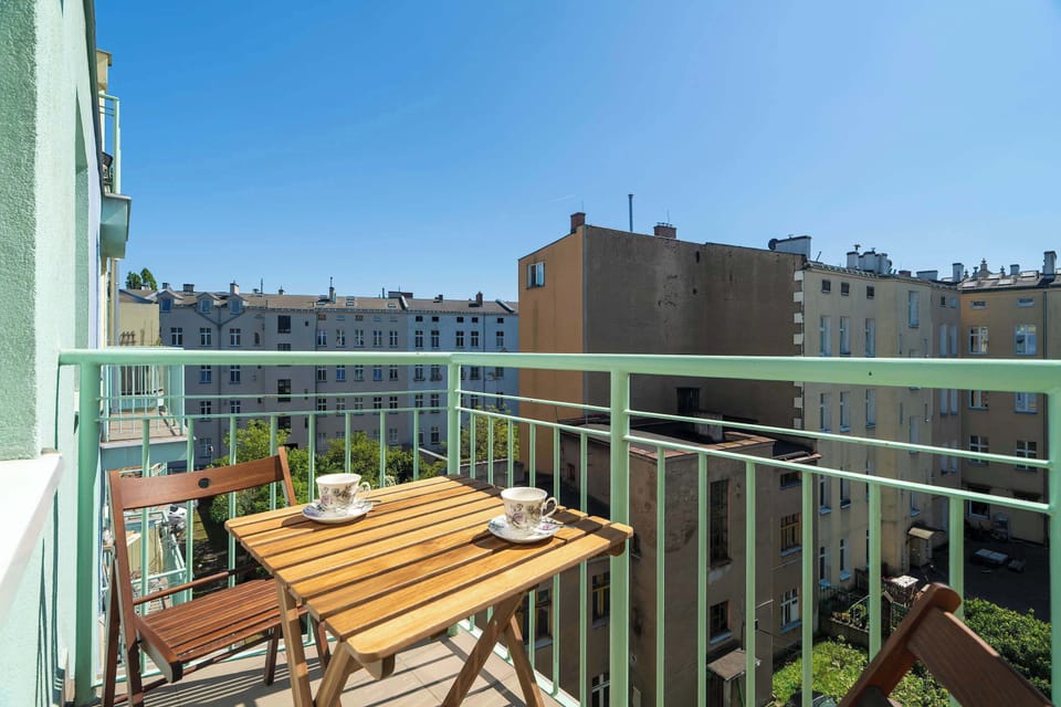 A balcony with a wooden table and chairs, overlooking a cityscape with buildings and green rooftops.