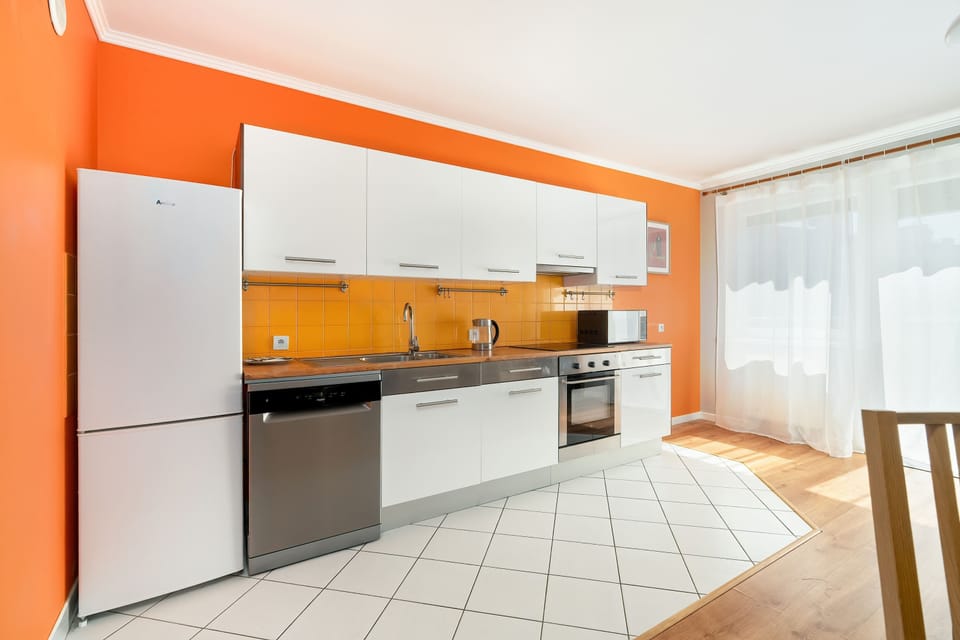 A modern kitchen with orange walls, white cabinetry, and tiled floors. The open design connects it to the living space.