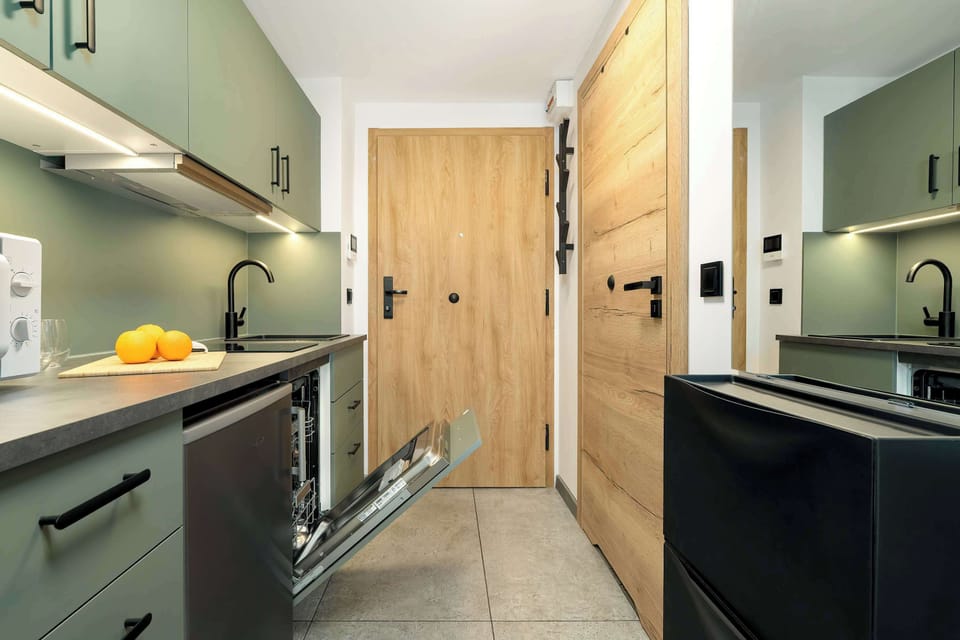 A modern kitchenette with green cabinets, a black sink faucet, and wooden accents, equipped with essential kitchen appliances including a dishwasher, and a cutting board with oranges, all presented in a neat and minimalist style.