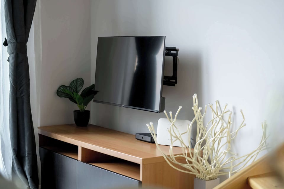 A modern living room with a wall-mounted TV above a wooden cabinet, which holds an elegant white branch-like decoration, a black pot with a green plant, and an electronic device, all set against light-colored walls and curtains.