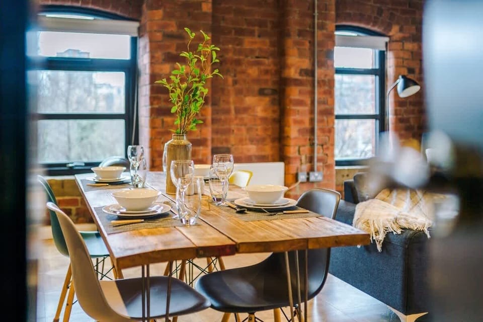 Stylish industrial-style dining area featuring a rustic wooden table set for four, complemented by modern chairs. Surrounded by exposed brick walls and large windows, the space is bright and inviting—perfect for shared meals or working remotely.