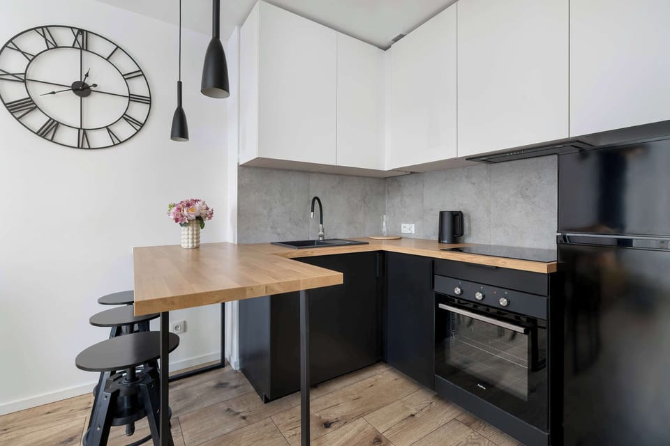 Small kitchen corner with electric stove, black and white cabinets and wall clock. Designed efficiently for everyday use.