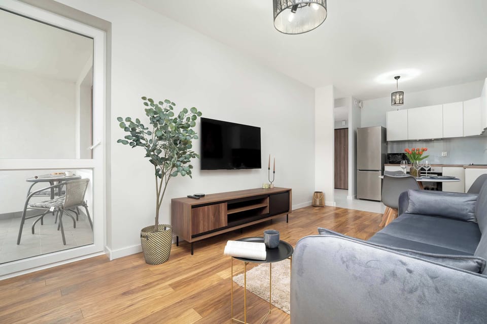 The modern living room combined with the kitchenette features wooden flooring, a gray sofa, a side table with a book and cup, a large potted plant, and a wall-mounted TV above an elegant TV stand.