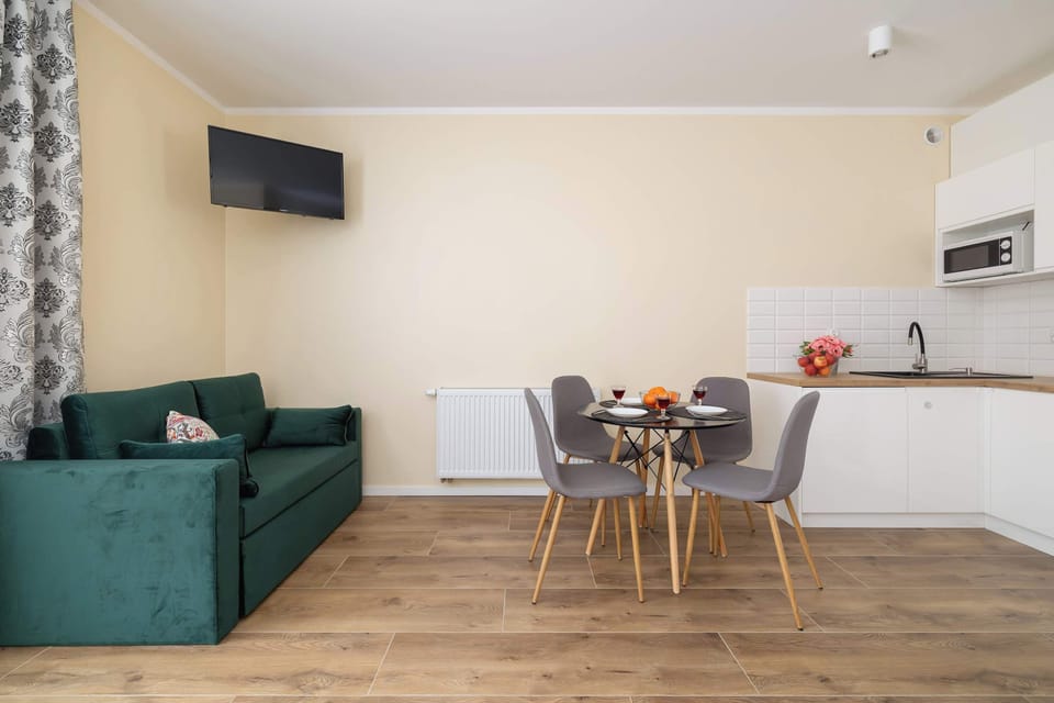 A spacious dining area with a TV mounted on the wall and a green sofa in the background. The wooden flooring enhances the warm and cozy ambiance.
