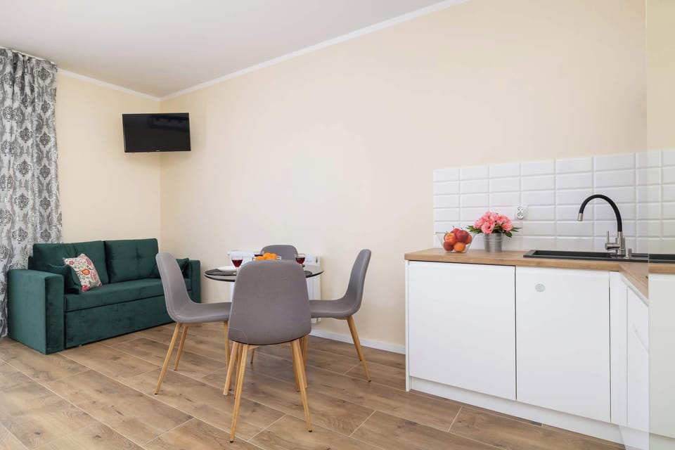 A modern living area with an open kitchen, dining table, and a green sofa. The room is well-lit with warm lighting and wooden flooring.