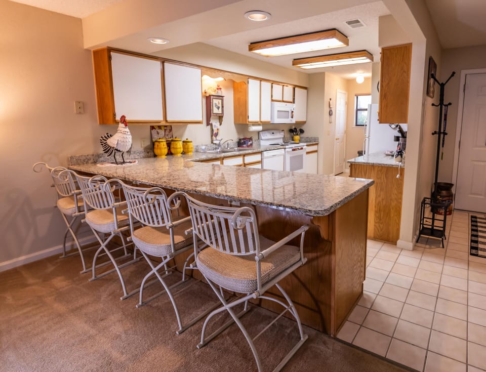 Granite countertop breakfast bar with cushioned wrought iron stools separates carpet and tile flooring in kitchen.