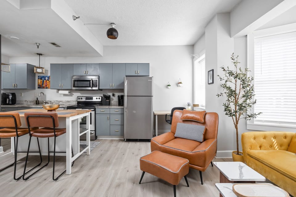 Designed for both style and function, this open-concept space features a mid-century chair, a cozy tufted couch, and a fully equipped kitchen—bringing comfort and character together in one seamless setting.
