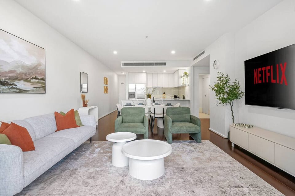 Stylish open plan living area with soft tones, modern seating, and a large TV for relaxed entertainment, flowing seamlessly into a bright dining space and kitchen.