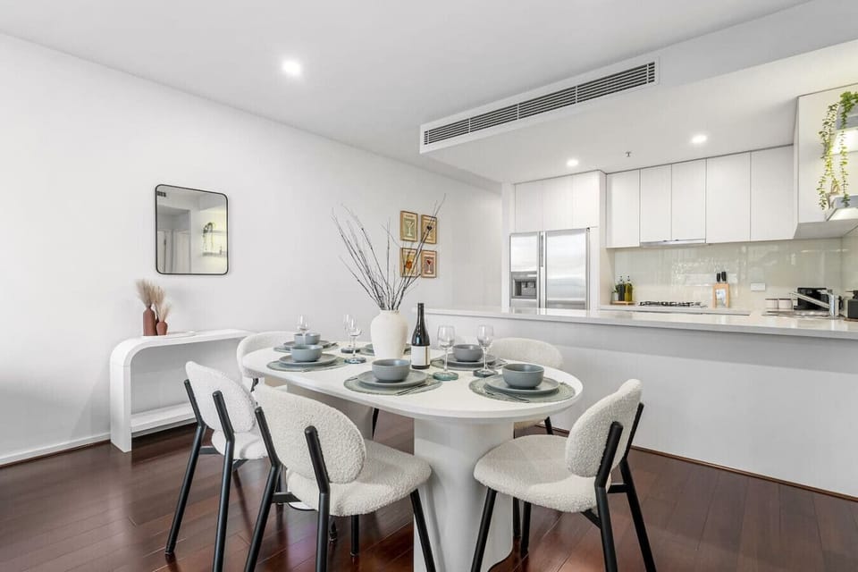 Bright dining area with a modern table setting, soft textured chairs, and a sleek white kitchen backdrop creating a clean and welcoming space for shared meals.