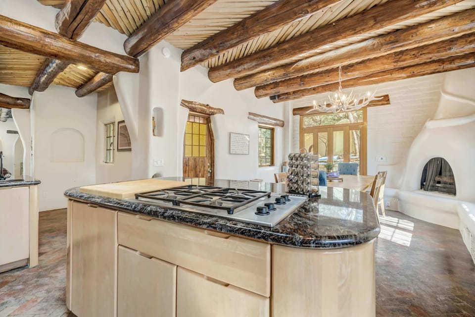 The central island features a gas cooktop and ample prep space beneath exposed log beams. The open layout connects seamlessly to the dining area and cozy fireplace beyond.
