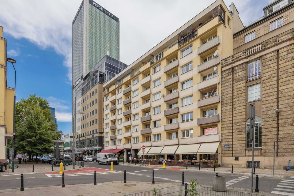 City street view with a modern skyscraper in the background, residential building with balconies in the foreground, pedestrian crossing, bollards, and trees on the left.