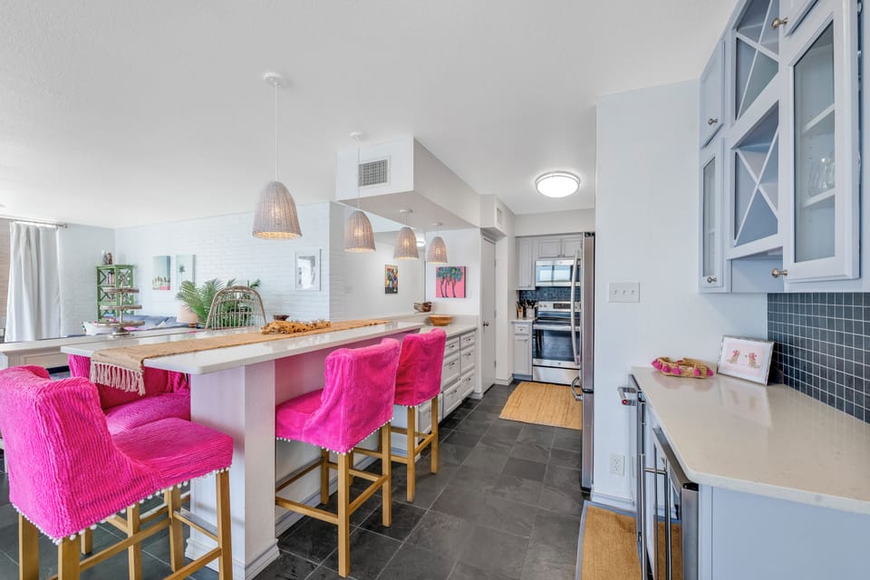Your bright kitchen awaits with vibrant pink seating and sleek appliances, perfect for creating memorable meals together.