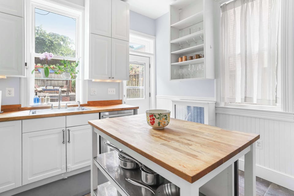 Kitchen that features center table with pans, kitchen sink, kitchen sink, kitchen cabinets, dishwasher, wine glasses on display, good lighting coming from window and a door leading to the patio.