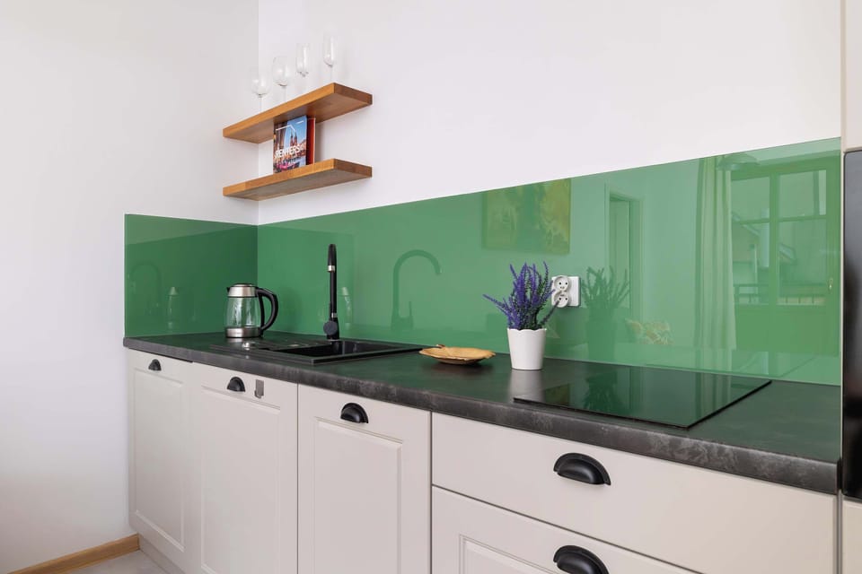A modern kitchenette with white cabinets, a black countertop, green backsplash tiles, and basic appliances such as an electric kettle and stovetop provides convenience during a short stay.