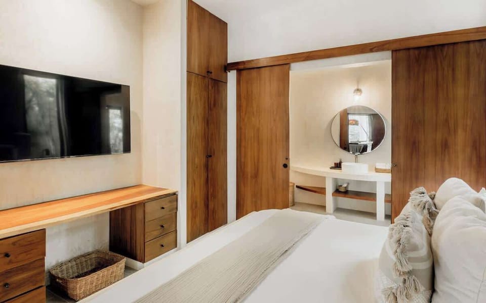 Bedroom with wall-mounted TV, dark wood dresser, round mirror, and vanity desk.