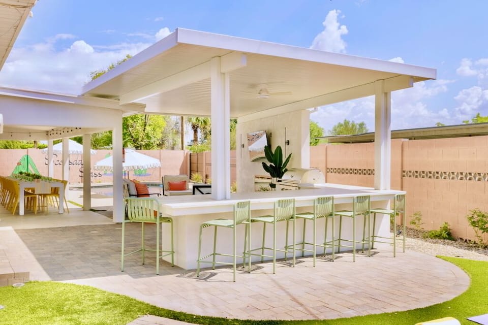 Resort-style backyard bar with outdoor kitchen, grill, TV, dining and shade.