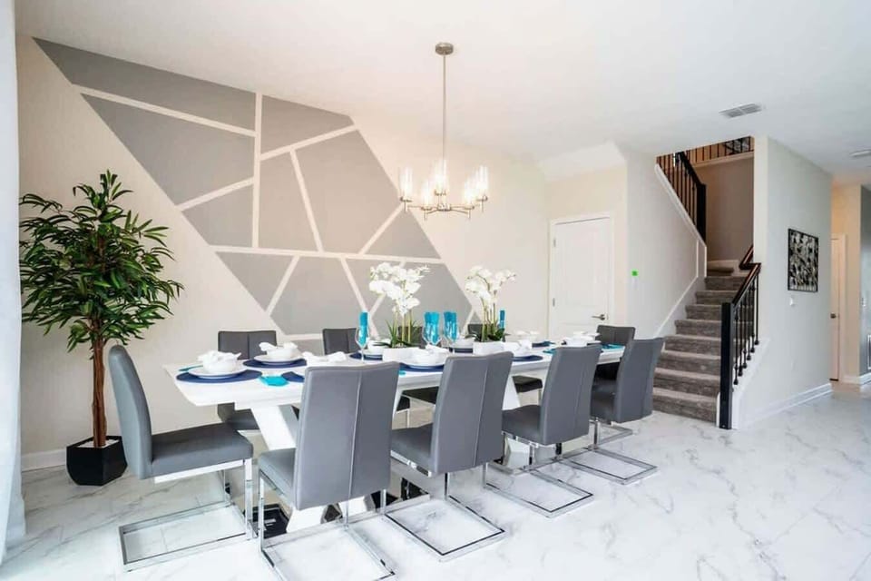Gather around the sleek white dining table for ten in this modern space, complete with a geometric accent wall, chic gray upholstered chairs, a statement chandelier, and marble-look floors-where every meal feels special!